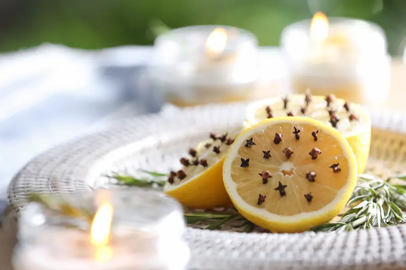 Halved lemons studded with whole cloves on a white woven plate, surrounded by fresh rosemary sprigs and blurred burning candles in the background.