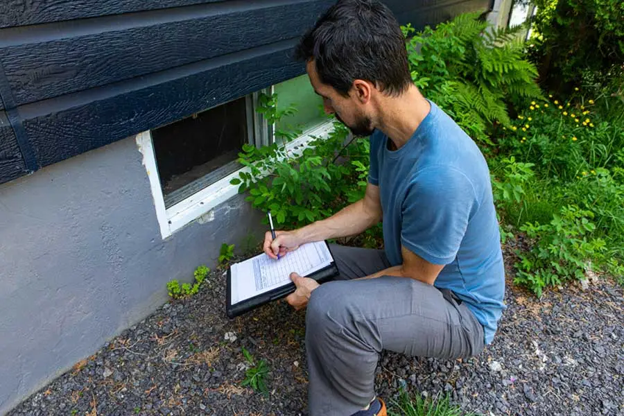 A man filling out a Wood Destroying Insect (WDI) inspection report on a clipboard while squatting outside a house.